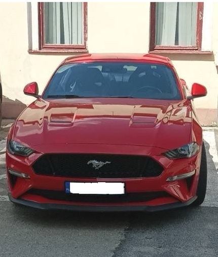 Czerwony samochód sportowy zaparkowany przed budynkiem z oknami osłoniętymi zasłonami.