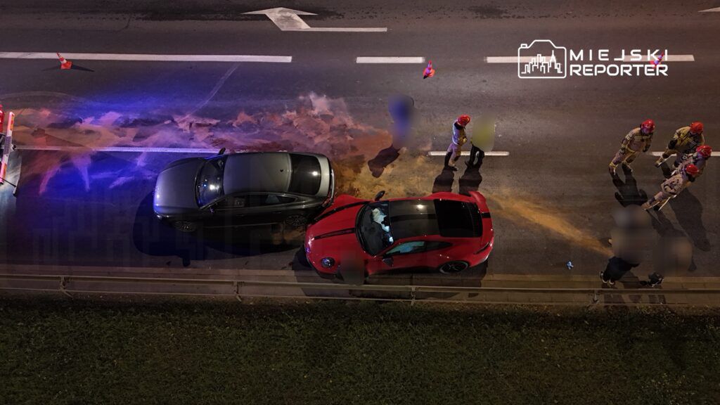 Dwa uszkodzone samochody, czarny i czerwony, zatrzymane na drodze, obok grupy strażaków w hełmach.