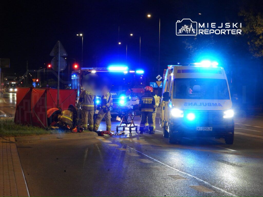 Grupa strażaków udziela pomocy osobie poszkodowanej na drodze, obok ambulansu i wozu strażackiego w nocy.