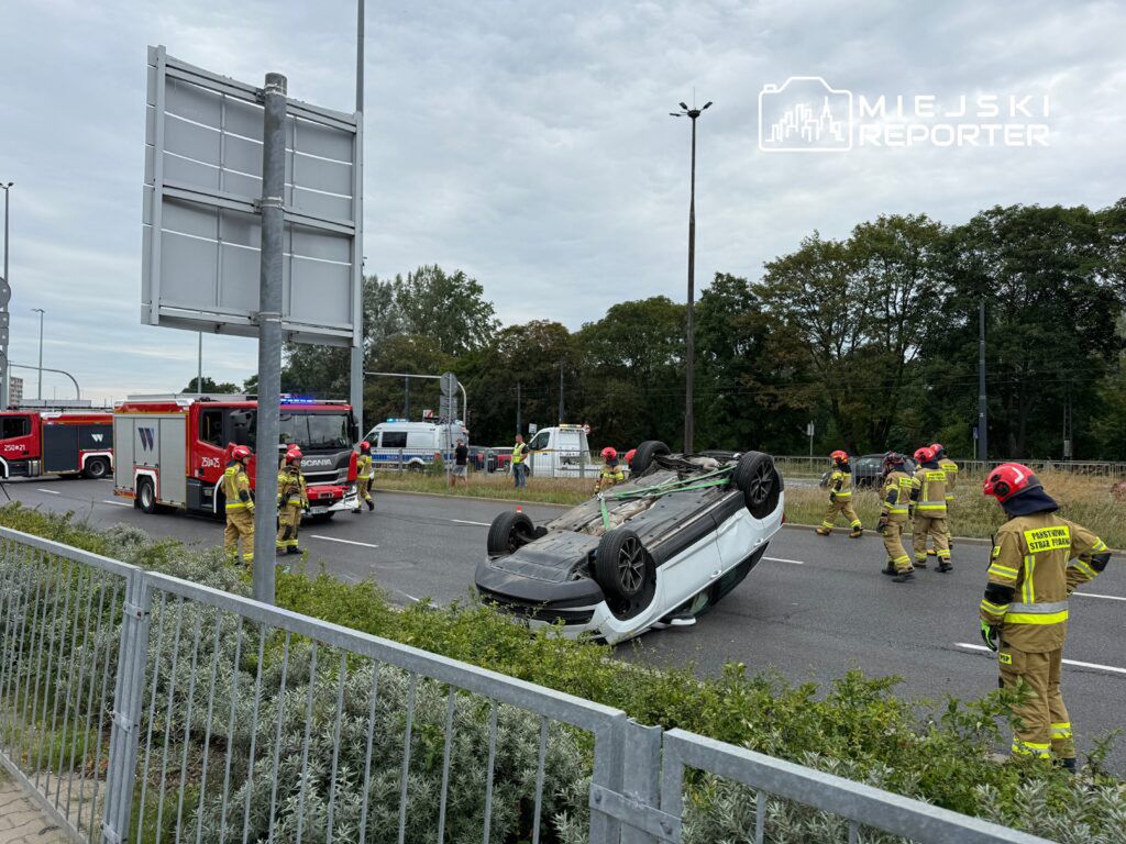 Na drodze leży przewrócony samochód, a w pobliżu grupa strażaków w hełmach ocenia sytuację.