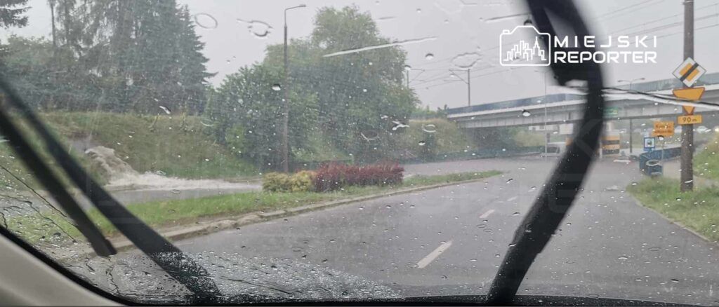 Intensywna ulewa z widocznymi kroplami deszczu na szybie samochodu, w tle zarośla i most.