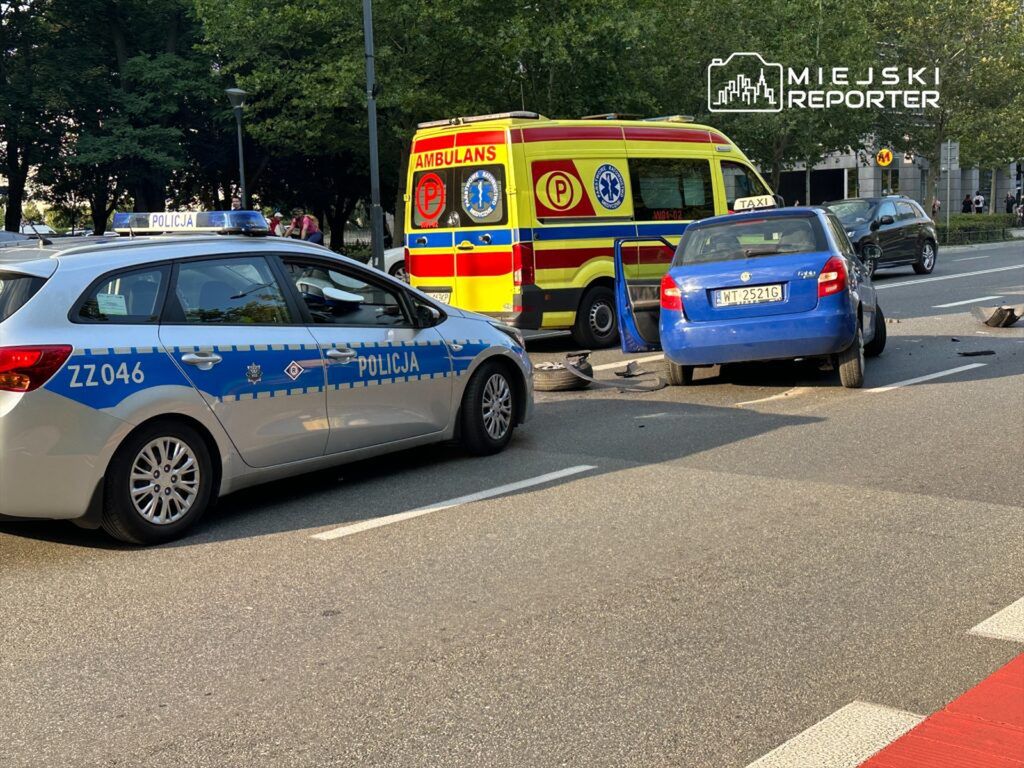 Nieoznakowany radiowóz policyjny oraz ambulans zatrzymane obok niebieskiego samochodu osobowego po wypadku drogowym.