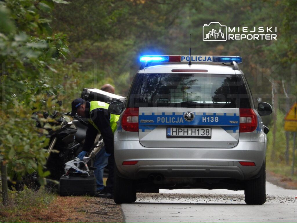 Funkcjonariusz policji sprawdzający motocykl przy drodze w lesie, obok radiowozu z włączonymi sygnałami świetlnymi.