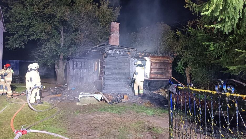 Strażacy gaszą pożar drewnianego budynku w nocy, otoczeni dymem i zniszczeniami na terenie posesji.
