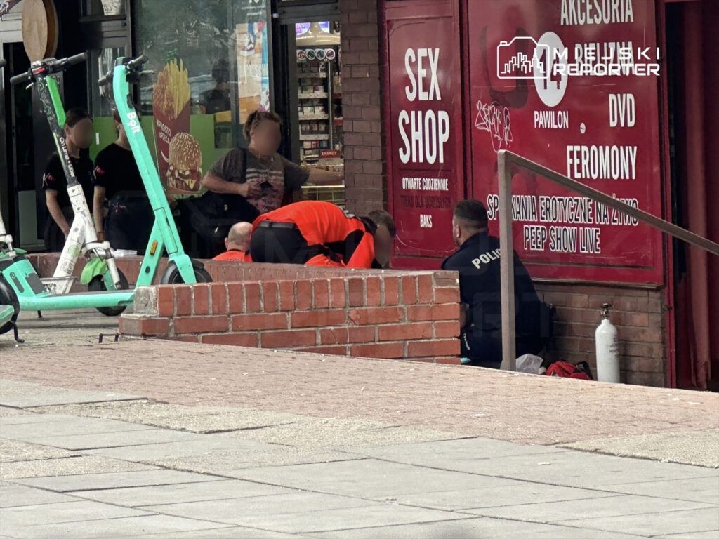 Grupa osób w pobliżu sex shopu, w tym policjant, udziela pomocy mężczyźnie leżącemu na ziemi.
