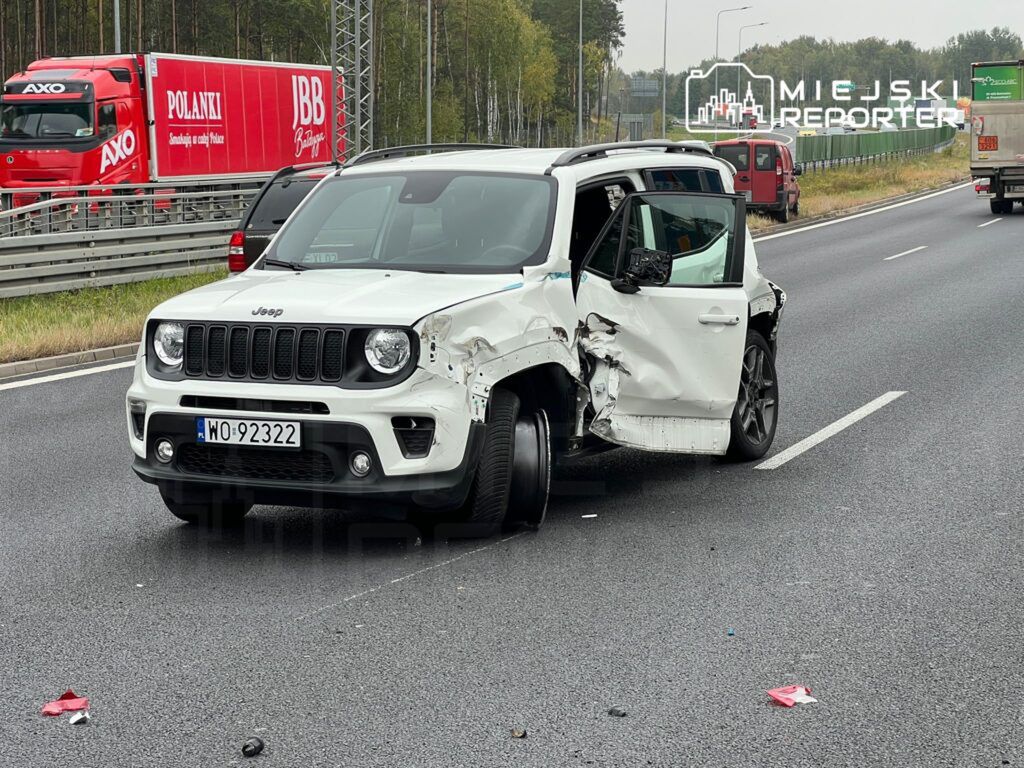 Uszkodzony biały SUV marki Jeep stoi na autostradzie z otwartymi drzwiami, obok widać ciężarówkę w ruchu.