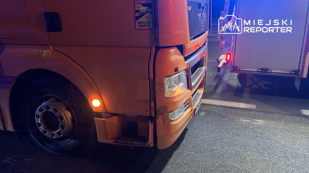 Pomarańczowy ciężarówka MAN z uszkodzoną przednią częścią stoi na drodze, obok widoczny pojazd ratunkowy.