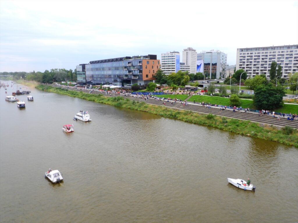 Wzdłuż rzeki pływają łodzie, a na bulwarze spacerują ludzie, w tle nowoczesne biurowce.