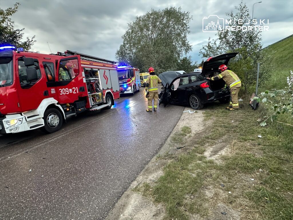 Strażacy w mundurach ratunkowych sprawdzają uszkodzony samochód osobowy przy drodze.