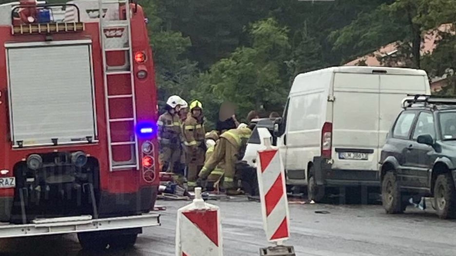 Grupa strażaków udziela pomocy poszkodowanemu na drodze, obok zaparkowanych pojazdów i oznaczeń robót drogowych.