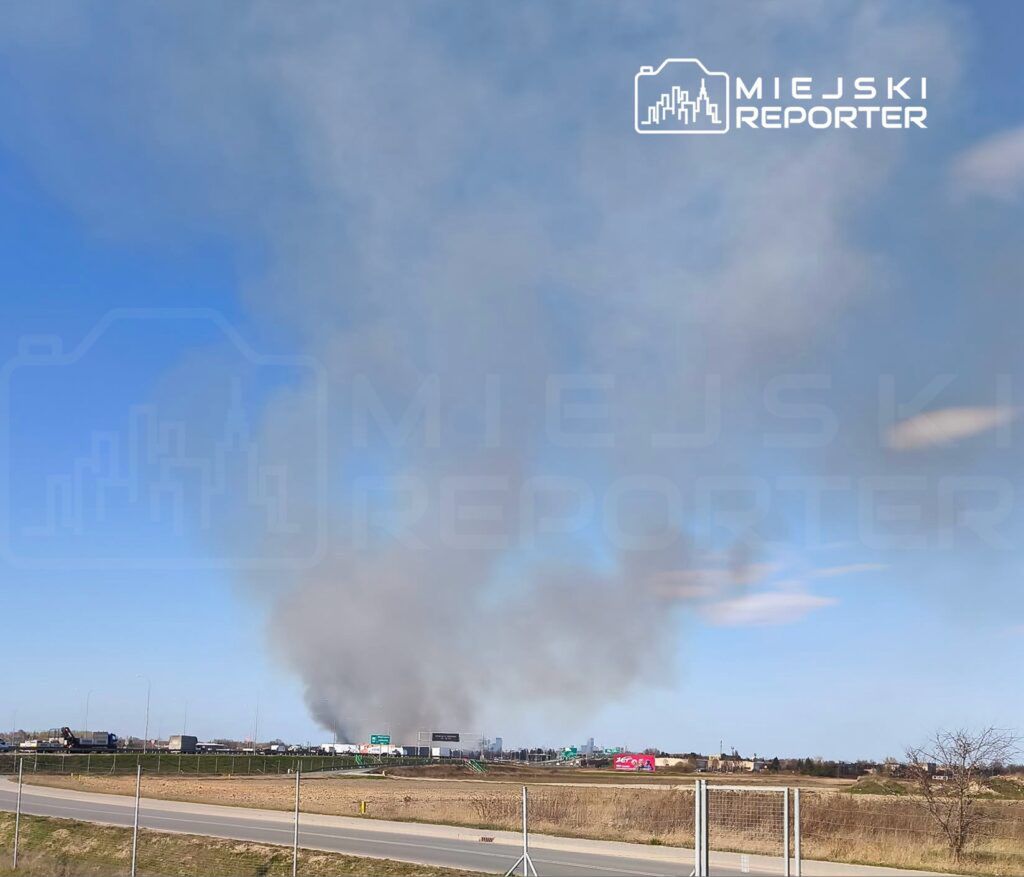 Gęsty słup dymu unoszący się nad terenem przemysłowym w słoneczny dzień. W tle widoczne znaki drogowe i budynki.