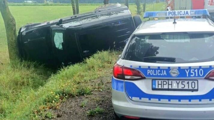Czarny samochód przewrócony na bok obok drogi, obok stoi radiowóz policyjny i wóz strażacki.
