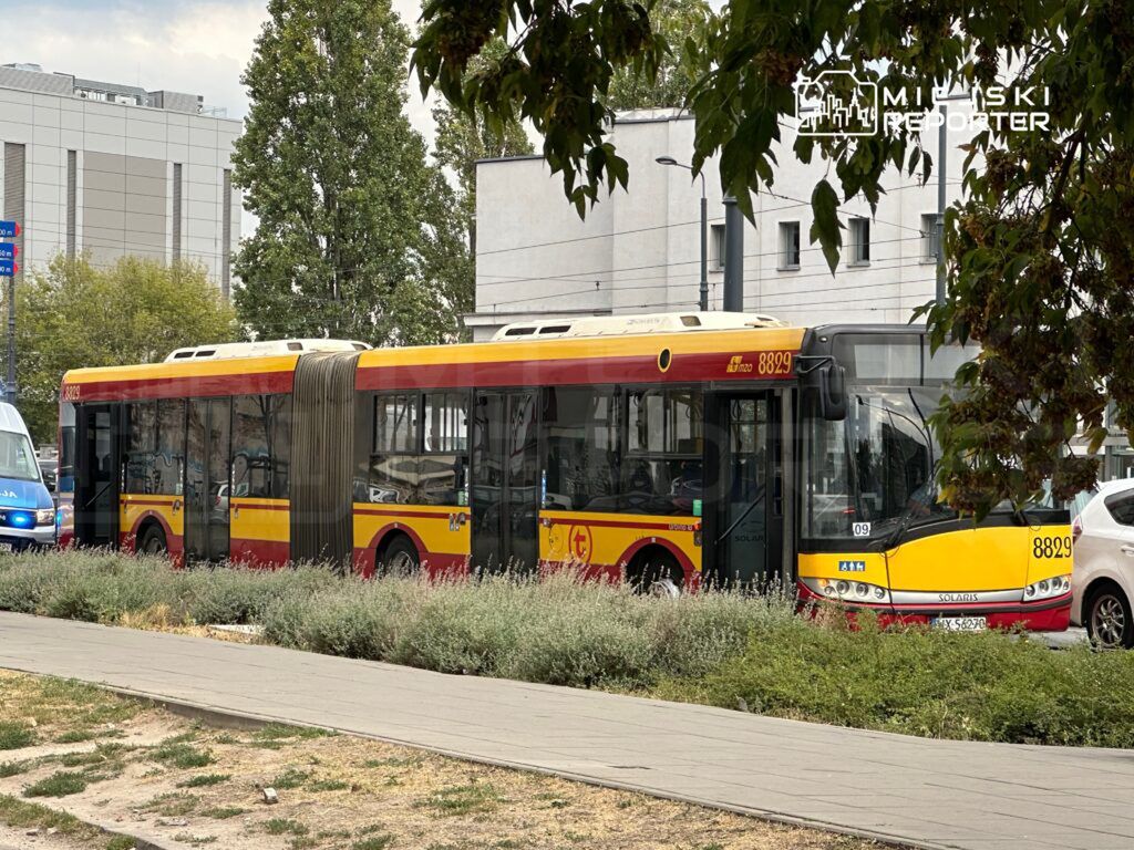 Autobus przegubowy w czerwono-żółtych barwach zatrzymany na przystanku, obok roślinności w miejskim otoczeniu.