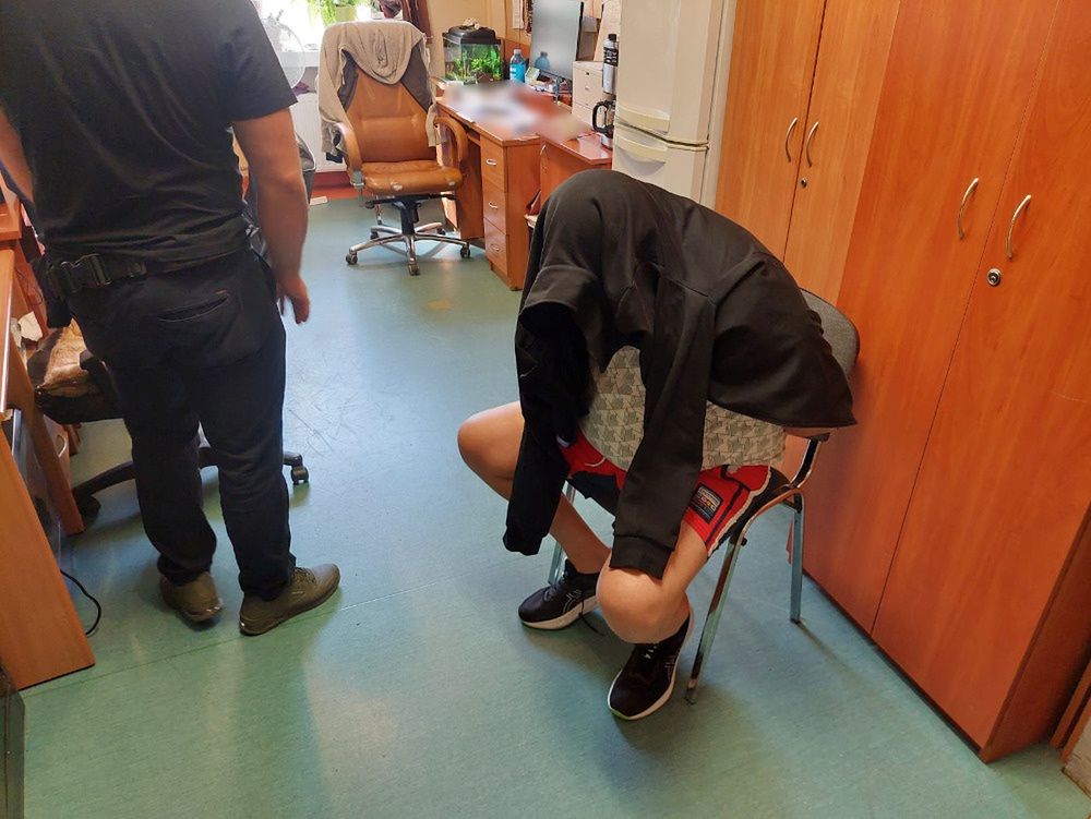 Mężczyzna w czarnej bluzie siedzi na krześle z głową opartą na kolanach, a obok stoi inny mężczyzna w ciemnych ubraniach.