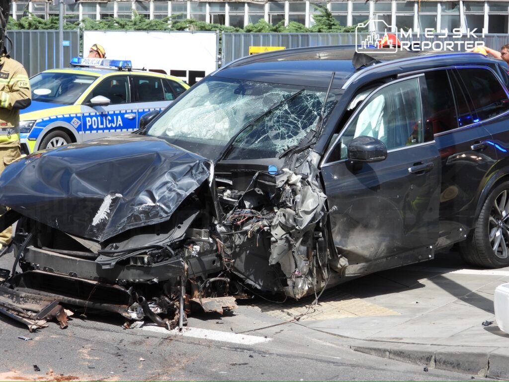 Rozbite czarne SUV z uszkodzoną przednią częścią, obok policyjny radiowóz oraz strażak w mundurze.