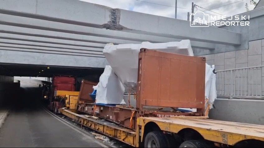 Ciężarówka z kontenerami przejeżdża pod niskim mostem, z widocznymi uszkodzeniami na betonowej konstrukcji.