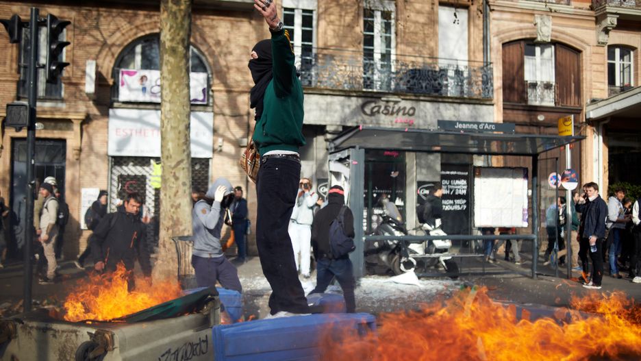 A man gestures on trash bins on fire during the riots. Scuffles happened between protesters and riot police during the protest as people reacted with anger to the interview of President Macron to justify the use the 49.3 article to adopt his pension reform without vote and the defeat of the motion of no confidence. Trade unions, political parties, youths, students took to the streets as evrywhere in France. More then 70,000 people protested in Toulouse. Polls show 67% french people say they want the stirkes to intensify the strikes and that 93% of workers oppose the pension reform. The retirement age would be raised to 64 years (for 43 years of continued work) and the pensions will be lower specially for women, 'short careers' and low wages people. Riot police sused dozens of tear gas canisters and a water cannon to disperse the crowd. Toulouse. France. March 23th 2023. (Photo by Alain Pitton/NurPhoto via Getty Images)