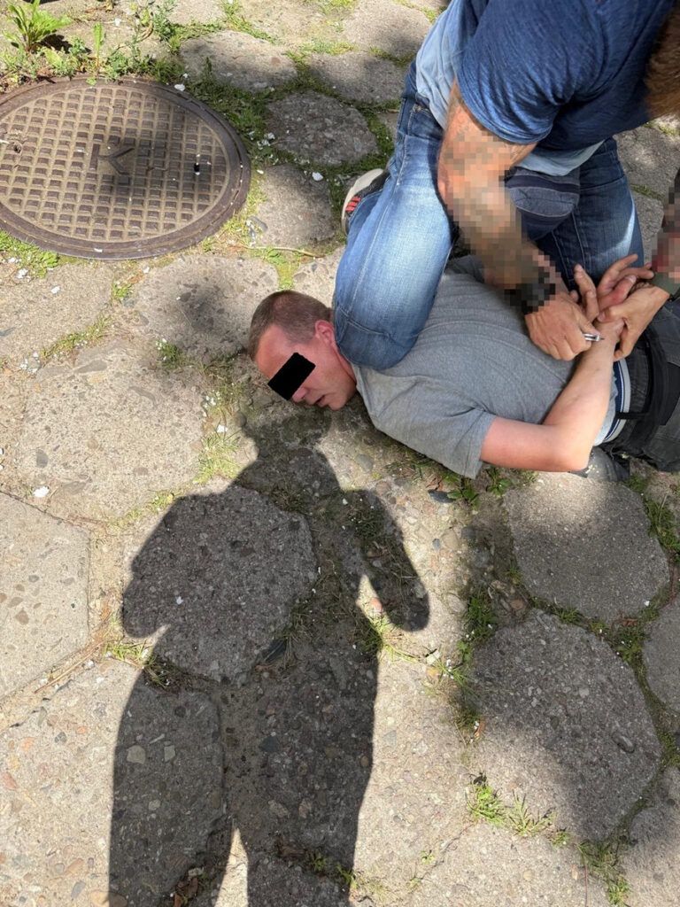 Mężczyzna unieruchomiony na ziemi przez innego mężczyznę, obaj na brukowanej nawierzchni, w tle cienie przechodniów.