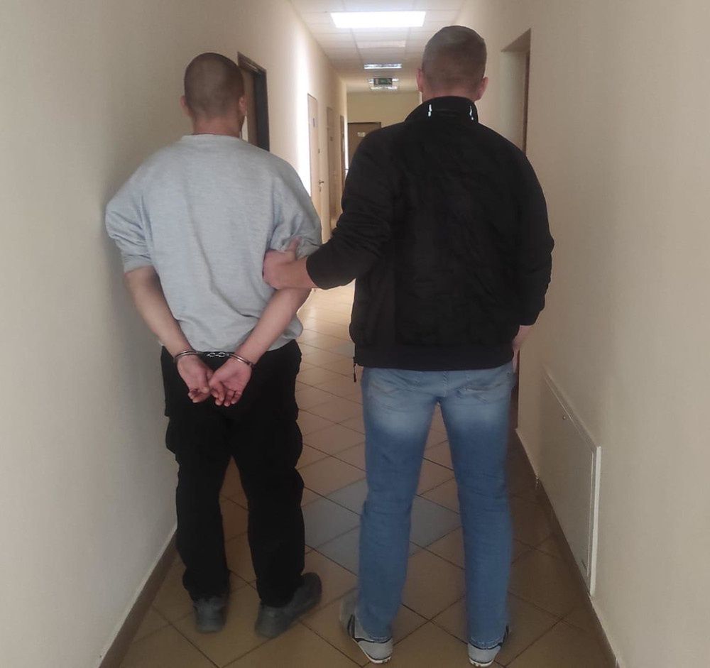 Mężczyzna w kajdankach prowadzony przez funkcjonariusza w korytarzu budynku.