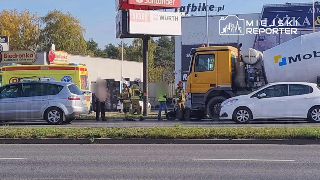 Grupa strażaków w mundurach pracuje przy wypadku drogowym obok cementowozu, w tle ambulans i sklep.