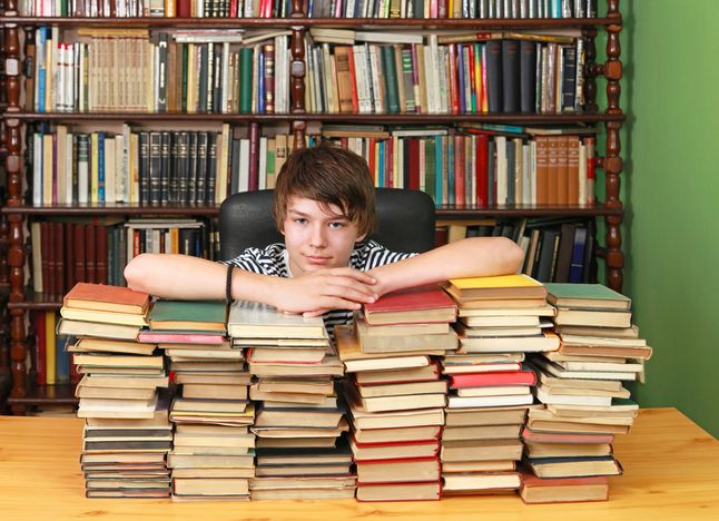 Young teenage boy with big pile of books in library