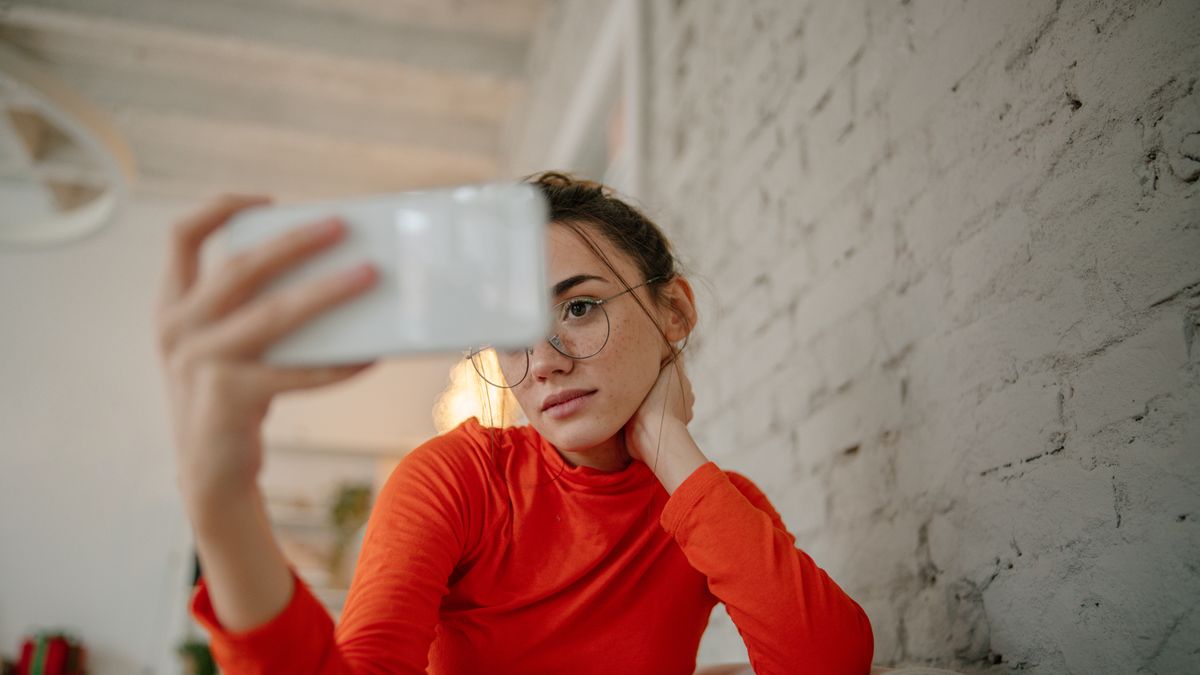 Photo of a young woman taking a self-portrait with a mobile phone while sitting in the living room of her apartment.