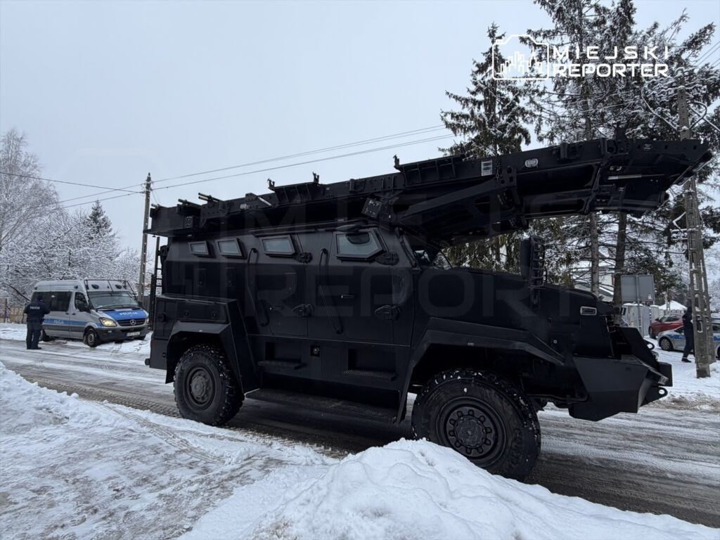 Czarny pojazd opancerzony z zamontowanym sprzętem na dachu stoi na zaśnieżonej ulicy obok policyjnego furgonu.