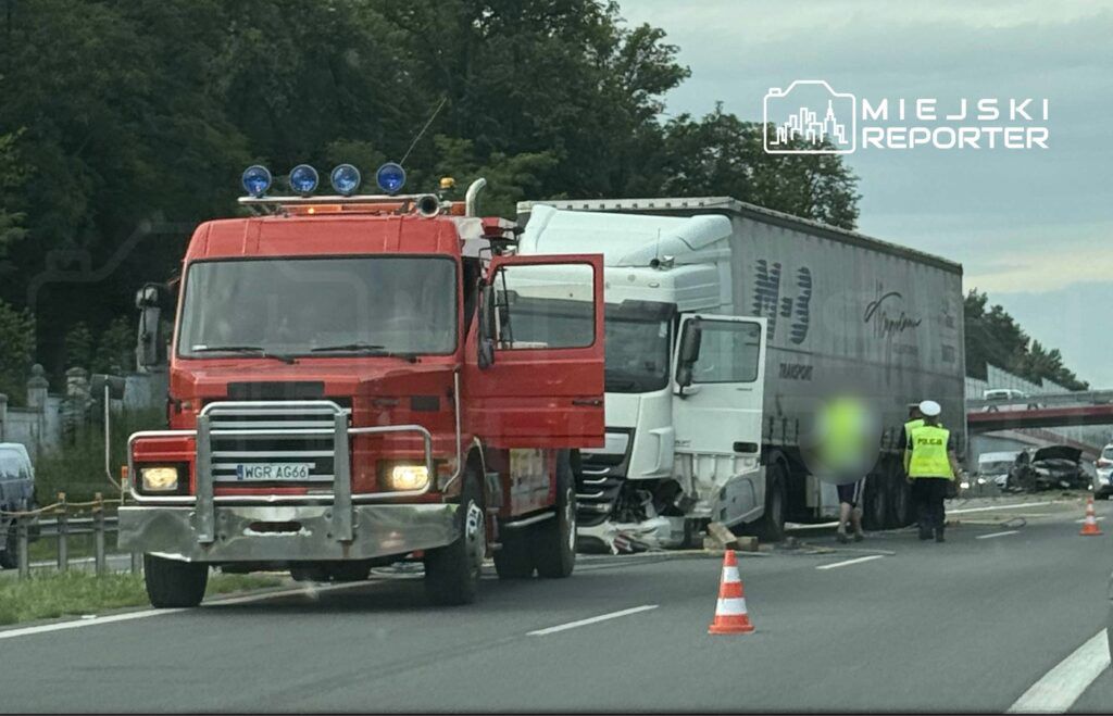 Czerwony wóz strażacki stoi obok zderzonej ciężarówki na drodze, w otoczeniu oznaczeń policyjnych i pachołków.