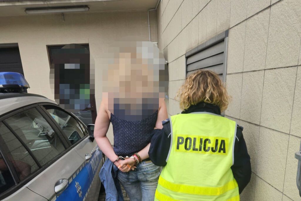 Kobieta z rękami związanymi na plecach stoi przy radiowozie policyjnym, a funkcjonariusz w odblaskowej kamizelce obserwuje sytuację.