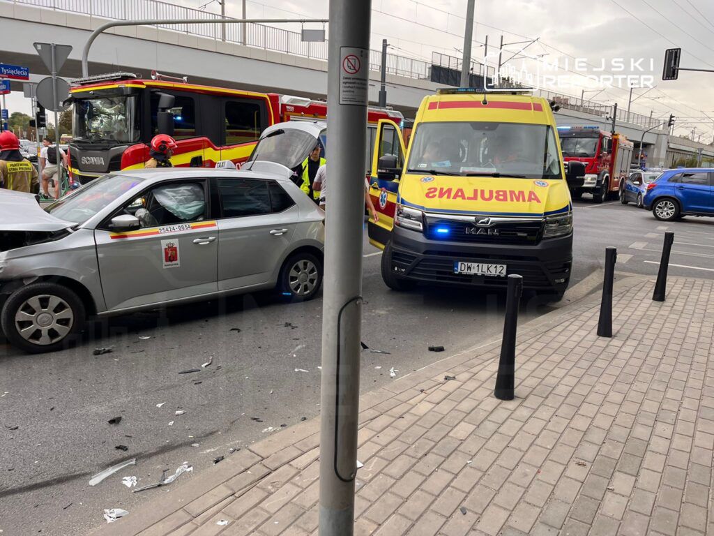 Wypadek drogowy z udziałem dwóch samochodów, w tym uszkodzonego pojazdu osobowego i karetki, na ruchliwej ulicy.