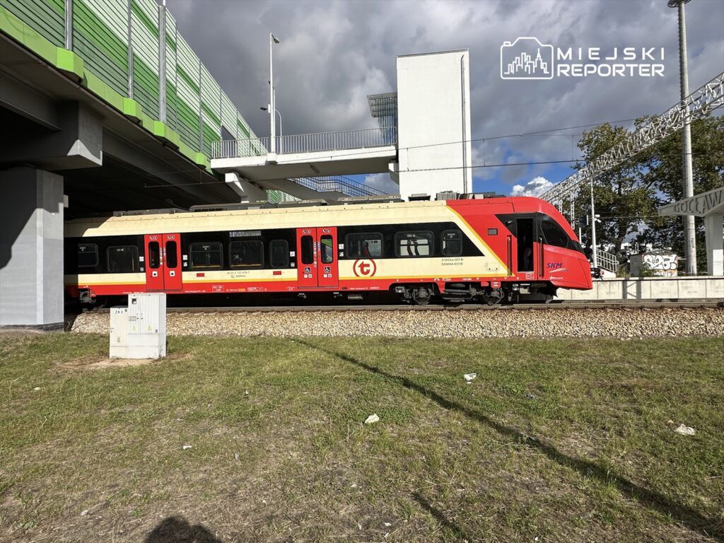 Pociąg SKM w czerwono-żółtych barwach przejeżdża pod nowoczesnym wiaduktem w miejskim otoczeniu.