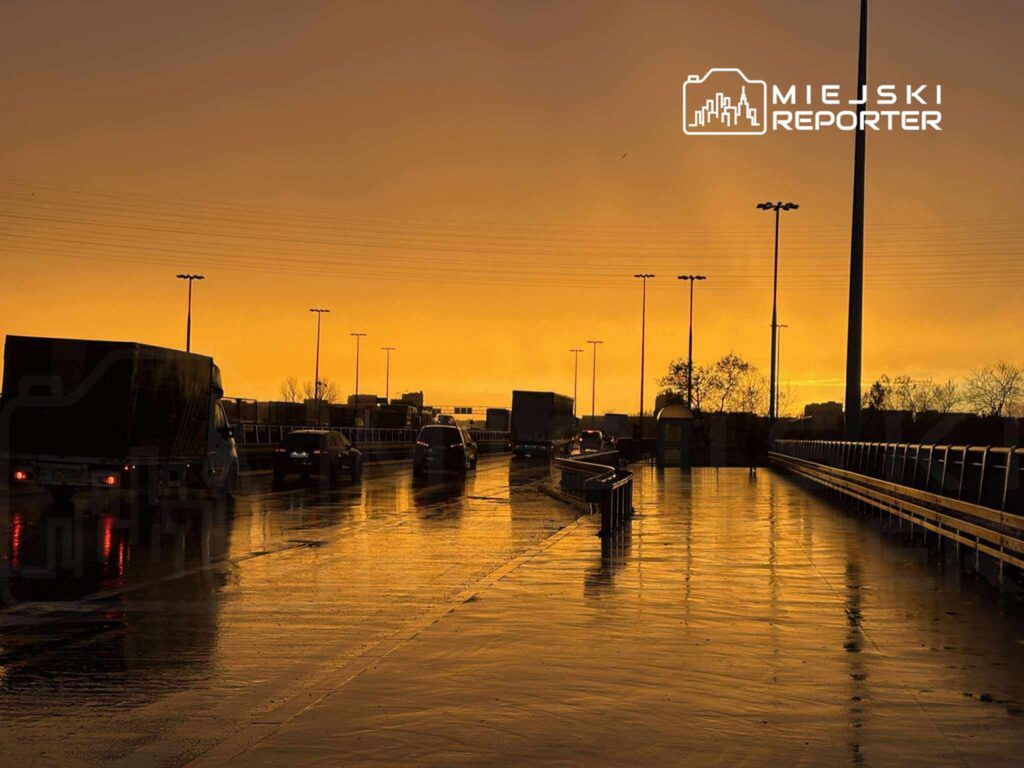 Samochody poruszają się po mokrej drodze w tle zachodzące słońce, oświetlające niebo na pomarańczowo.