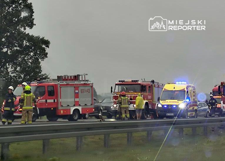Grupa strażaków oraz ambulans na poboczu drogi, w tle widoczne pojazdy służb ratunkowych. Deszczowa pogoda.