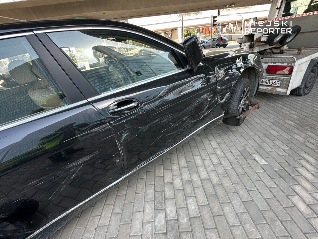 Czarny samochód osobowy z uszkodzeniami na prawym boku stoi na płycie chodnikowej obok lawety.