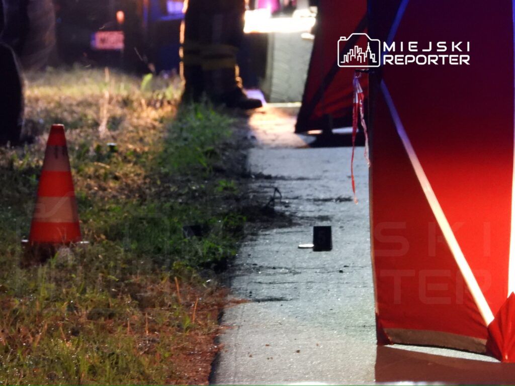 Na zdjęciu widoczna jest scena zdarzenia, z czerwonym namiotem zabezpieczającym oraz oznaczeniami na drodze. W tle widać pojazdy służb ratunkowych. W pobliżu znajduje się pomarańczowy pachołek, a na ziemi leżą różne przedmioty. To miejsce zostało zabezpieczone w związku z prowadzonymi czynnościami.