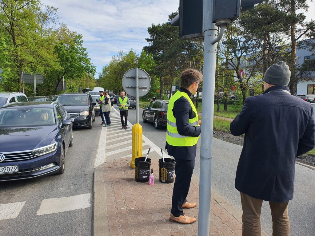 Grupa mężczyzn w kamizelkach odblaskowych prowadzi akcję informacyjną przy ruchliwej ulicy, obok zatrzymanych samochodów.
