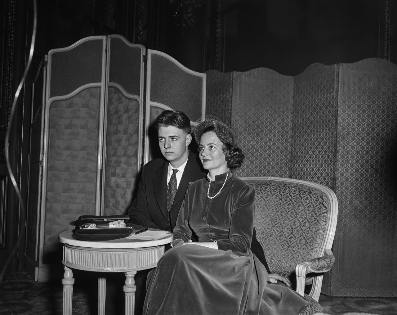 Albanian Royal Leka, Crown Prince of Albania, wearing a dark suit over a shirt and tie, and his mother, Queen Geraldine of Albania, who wears a velvet dress, a hat with the veil up, and a pearl necklace, sitting at a table with screens in the background, in London, England, 21st January 1957. (Photo by Evening Standard/Hulton Archive/Getty Images)