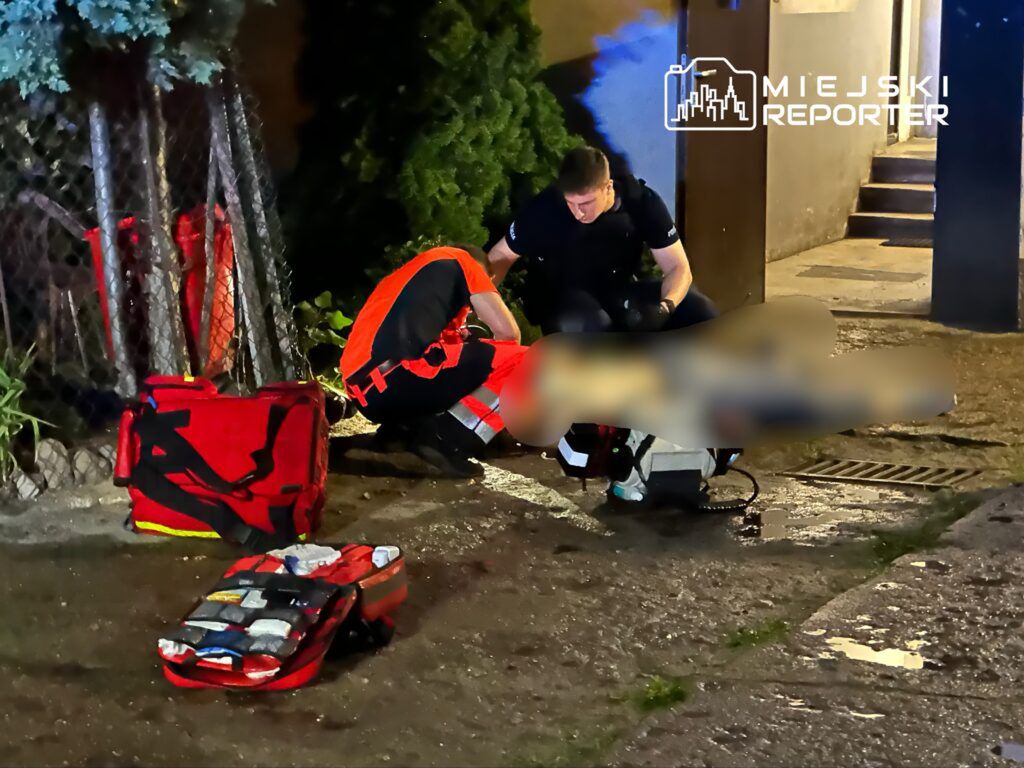 Ratownicy medyczni udzielają pomocy poszkodowanemu na chodniku, obok torów i ciemnej ściany budynku.