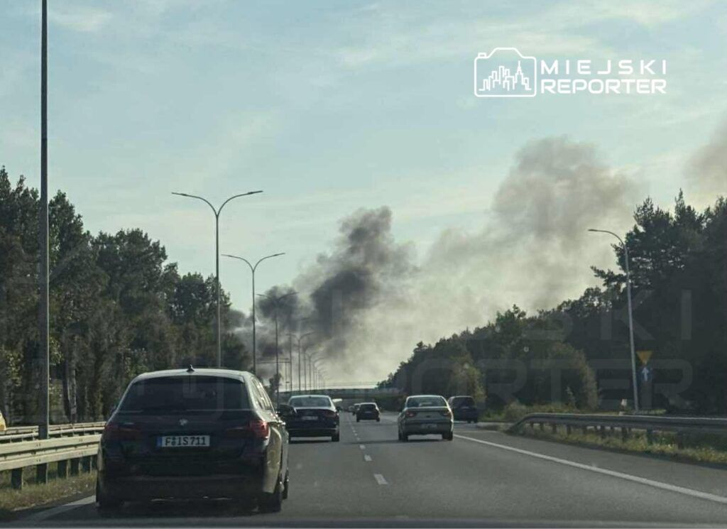 Na drodze widoczne są samochody, a w tle unosi się czarny dym z kierunku lasu.