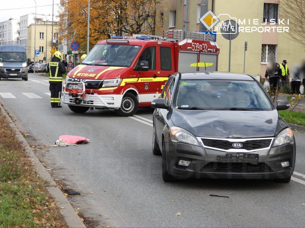 Strażacy w kamizelkach odblaskowych interweniują przy wypadku drogowym, obok leży różowa odzież na jezdni.