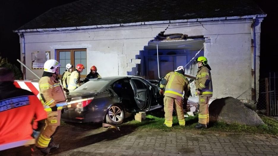 Grupa ratowników w mundurach pracuje przy uszkodzonym samochodzie, który wjechał w ścianę budynku.