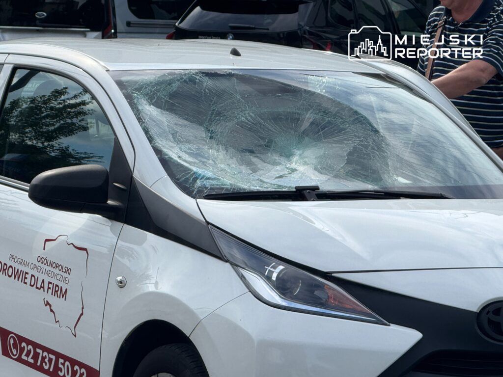 Rozbita szyba przednia białego samochodu osobowego zaparkowanego na parkingu, w tle widoczne inne pojazdy.