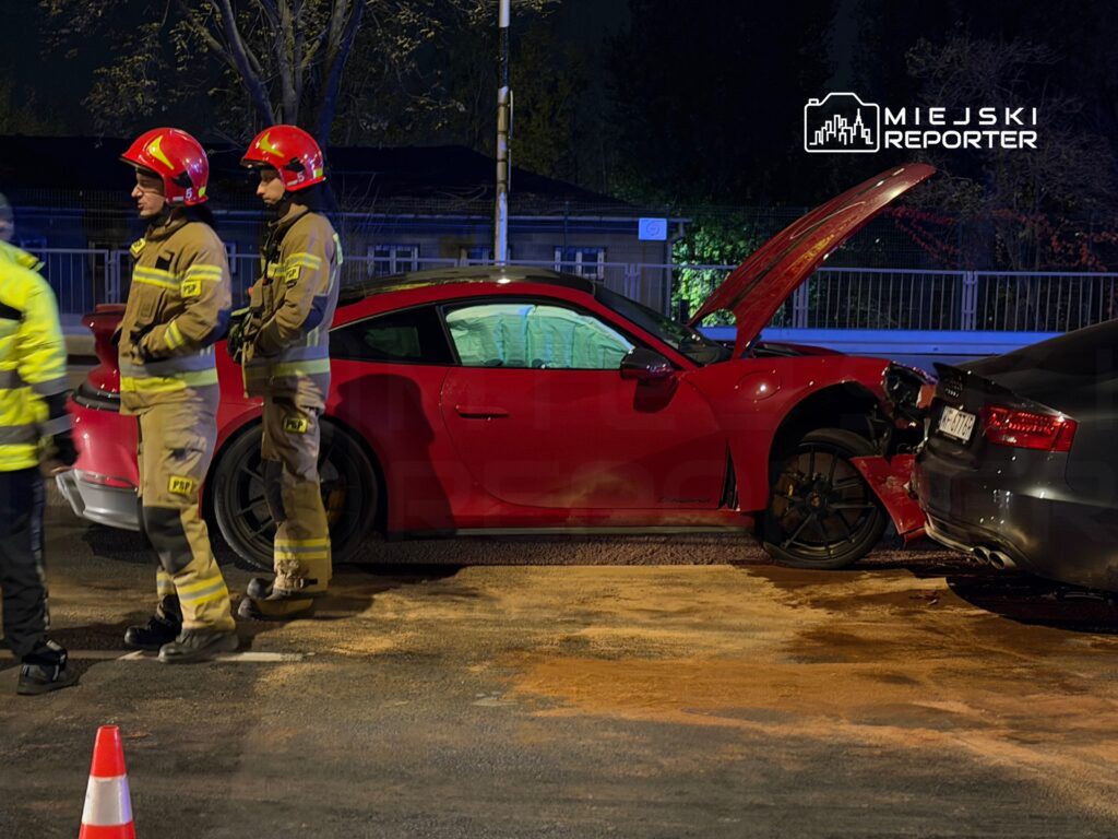 Dwóch strażaków w uniformach na drodze obok czerwonego sportowego auta z otwartą maską po zderzeniu z ciemnym samochodem.