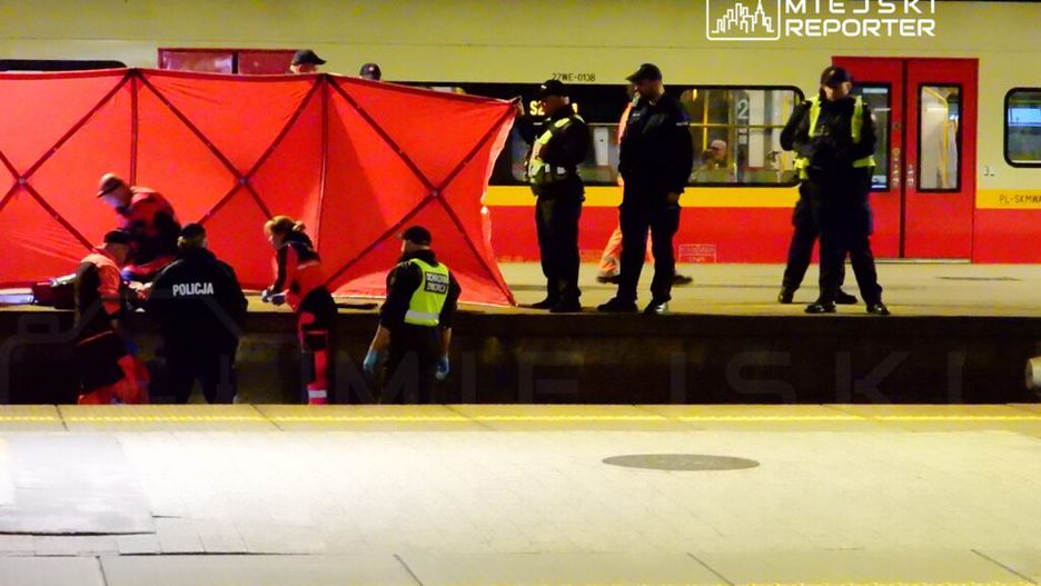 Grupa służb ratunkowych i policji pracuje przy peronie, zasłonięta czerwoną osłoną, w nocy.