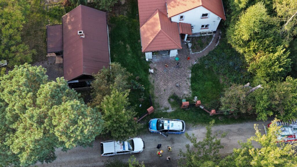 Na podjeździe do domu widać dwa pojazdy służb ratunkowych oraz kilka osób w mundurach, rozmawiających obok.