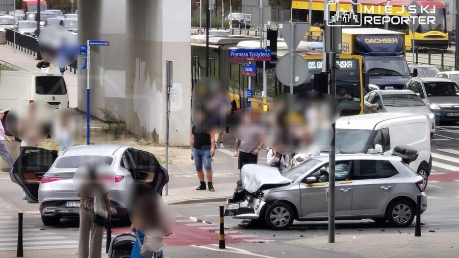 Zderzenie dwóch samochodów na ruchliwej ulicy, obok grupy osób obserwujących zdarzenie. W tle widoczne przystanki komunikacji miejskiej.