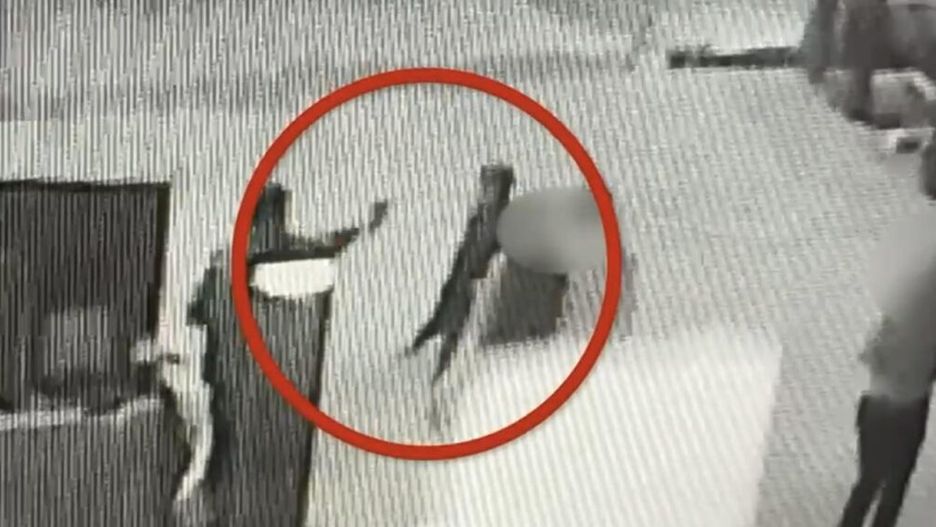 Osoba w ciemnym ubraniu trzyma przedmiot w ręku, stojąc w zamkniętym pomieszczeniu z widocznymi innymi osobami.