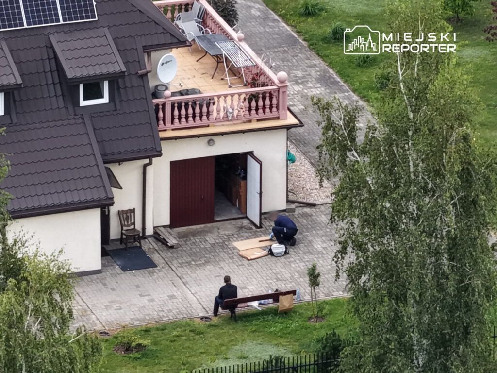 Mężczyzna w garniturze siedzi na ławce przed domem, podczas gdy inny mężczyzna pracuje przy materiałach budowlanych.