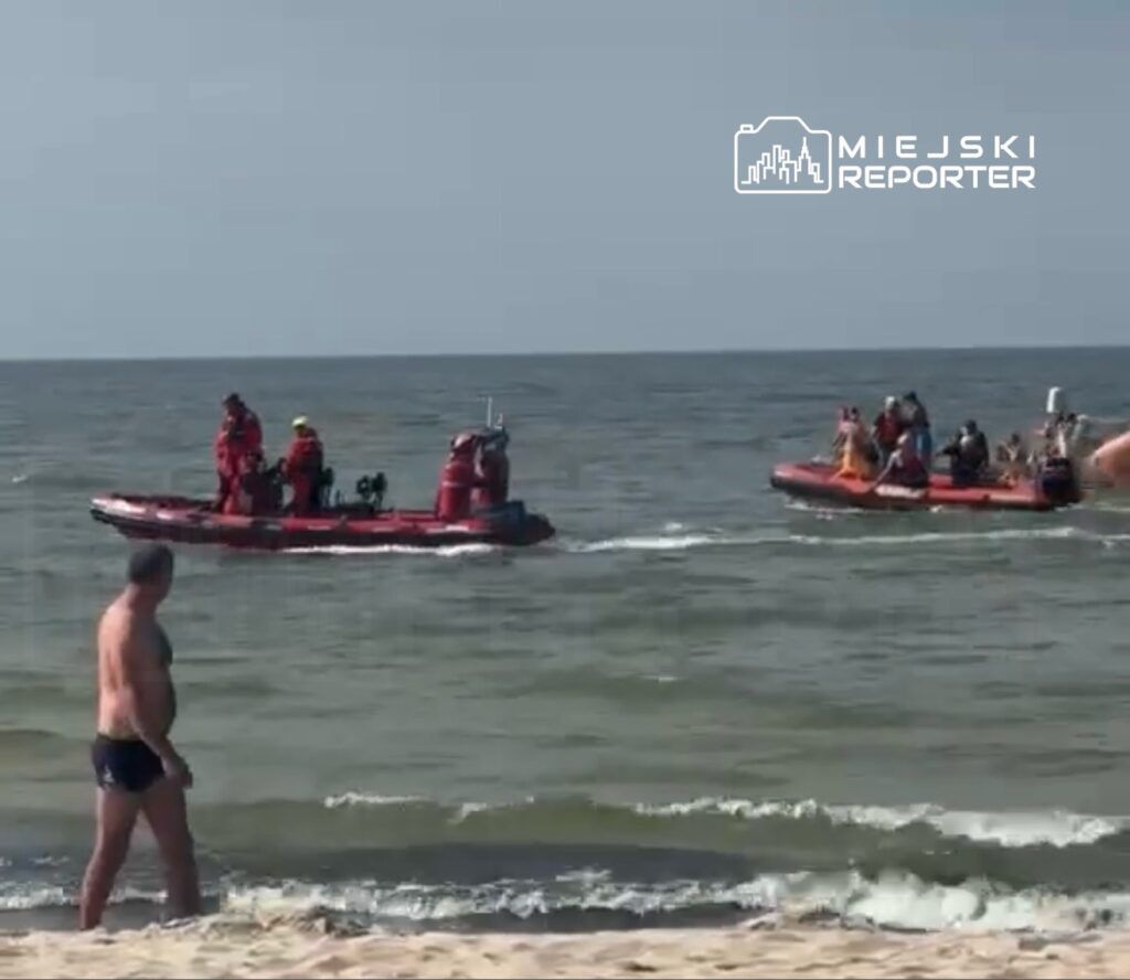 Mężczyzna w kąpielówkach spacerujący po plaży, obok przepływające łodzie ratunkowe z ratownikami.