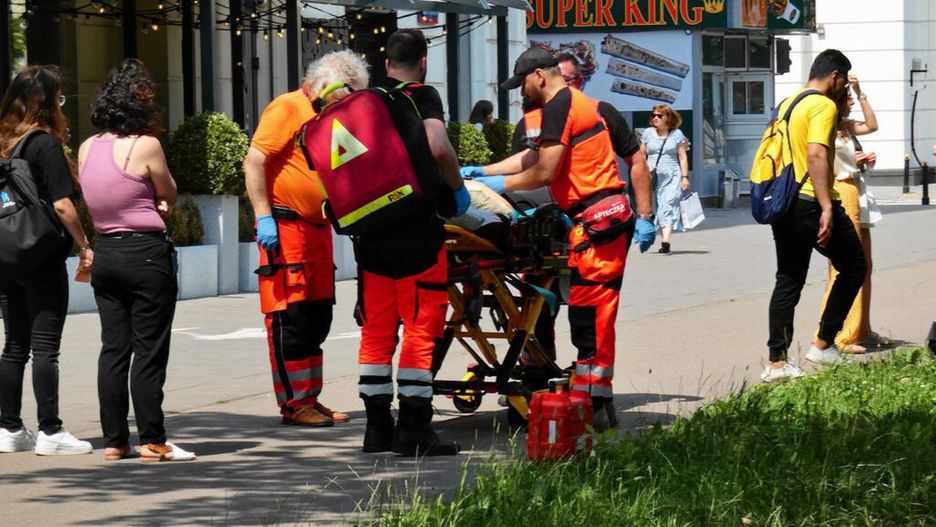 Zespół ratowników medycznych udziela pomocy osobie na noszach na ruchliwej ulicy. W tle przechodnie i lokalne sklepy.
